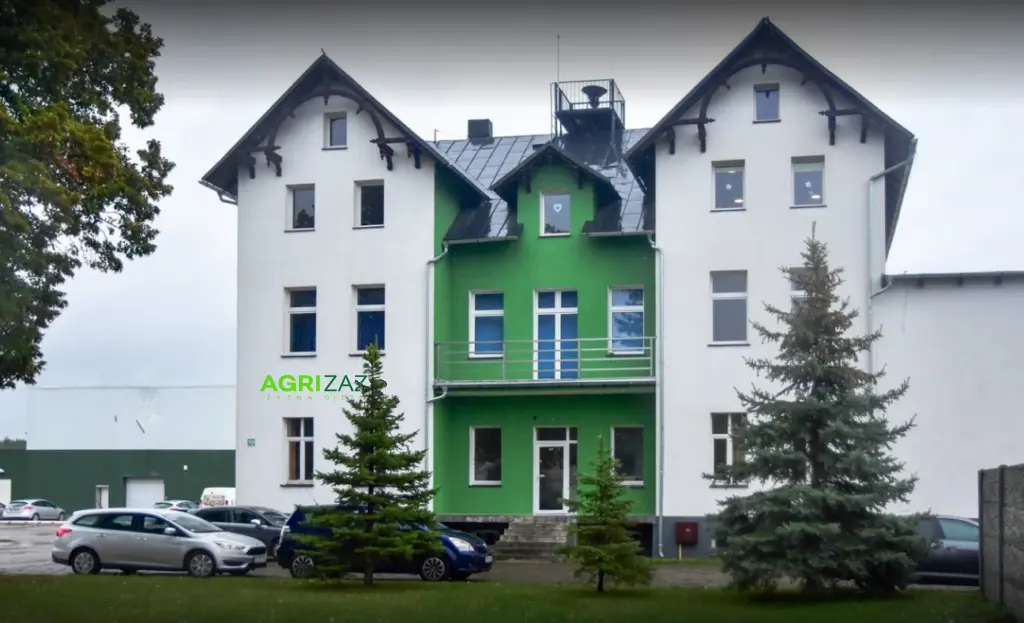 Trzypiętrowy budynek firmy AGRIZAZ o białej i zielonej elewacji, z parkingiem dla samochodów i rosnącymi przed nim drzewami iglastymi
